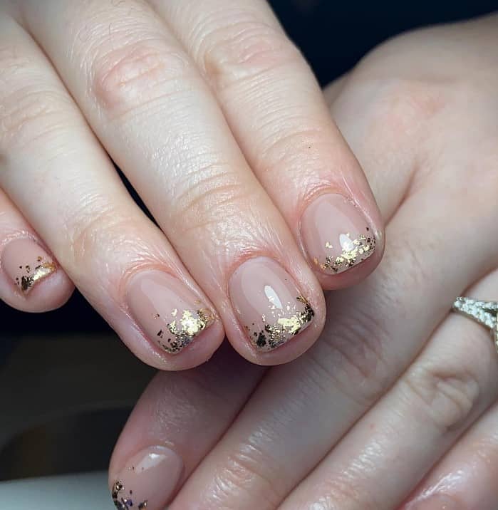 A closeup of a woman's fingernails with a glossy nude nail polish base that has gold foil French tips
