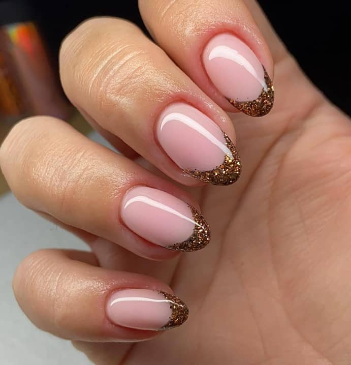 A closeup of a woman's medium-length fingernails with a glossy peach base that has dark gold glitter tips