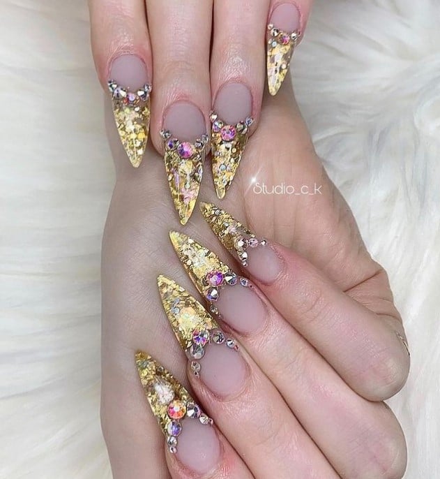 A closeup of a woman's long stiletto-shaped nails with a nude nail polish base that has French tips made of gold sequins with rhinestone borders