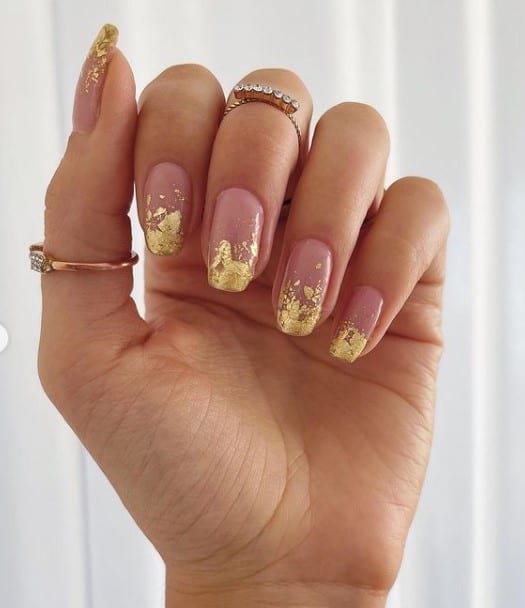 A woman's fingernails with a nude nail polish base that has gold foil French manicure with a subtle ombré effect