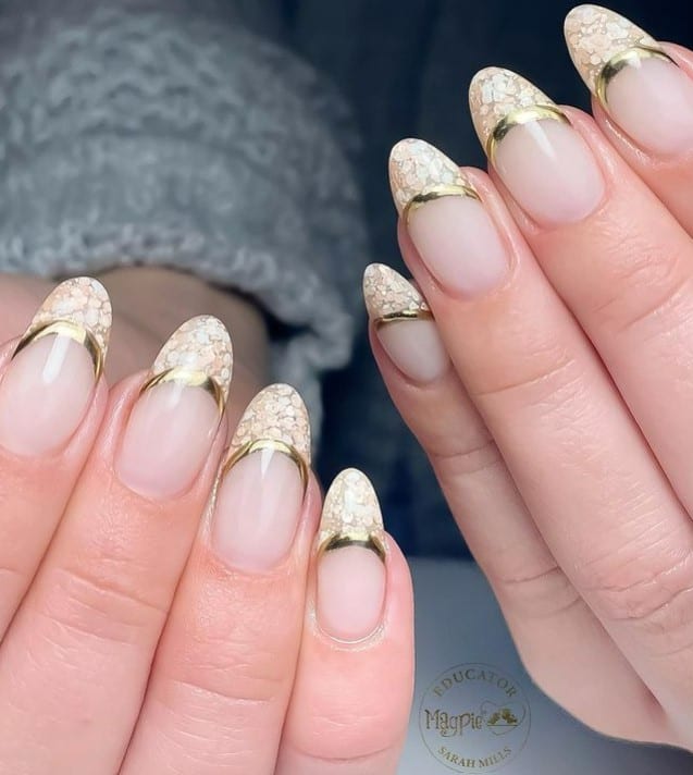 A woman's fingernails with a nude nail polish base that has French tips in chunky gold glitter and bold outlines in solid gold