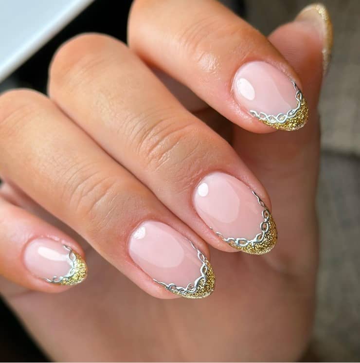 A closeup of a woman's fingernails with a glossy nude nail polish that has sparkling gold glitter French tips with chrome silver chains outlining each nail