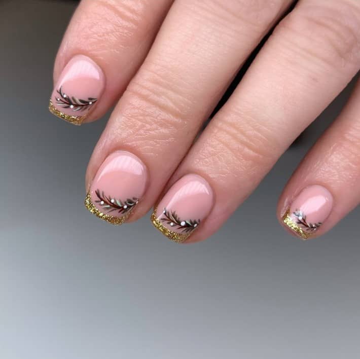 A closeup of a woman's fingernails with a glossy nude nail polish base that has gold French tips and festive green leaves
