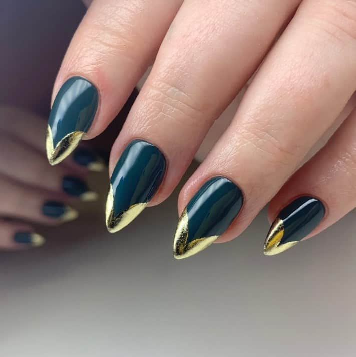 A closeup of a woman's almond-shaped fingernails with a deep bluish-green nail polish that has elegant heart-shaped French tips in luxurious gold