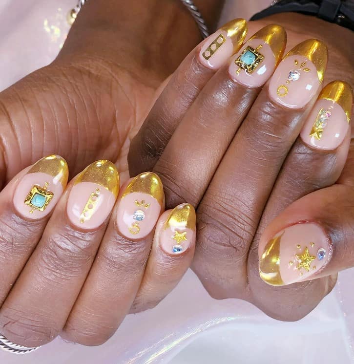 A closeup of a woman's fingernails with a peach nail polish base that has gold French tips, gold ornaments like turquoise-and-gold charms, gold stars, and round gems