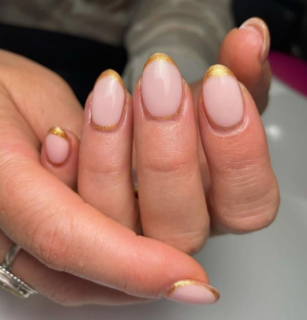 A closeup of a woman's fingernails with a nude nail polish base that has French tips in gleaming gold glitter highlighted with matching gold cuffs