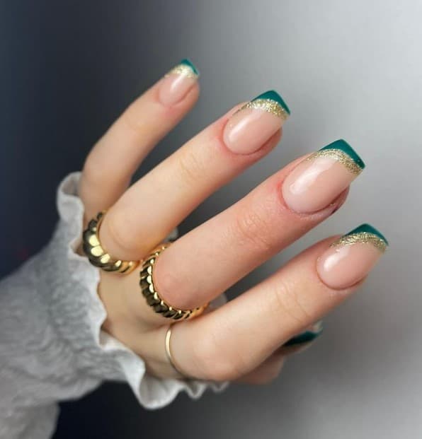 A closeup of a woman's fingernails with a a clear base that has deep green side French tips accented with shimmering gold borders