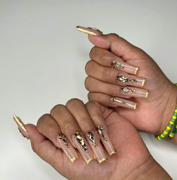 A closeup of a woman's fingernails with a peach nail polish base that has gold French tips and stunning gold rhinestones in different sizes on each nail