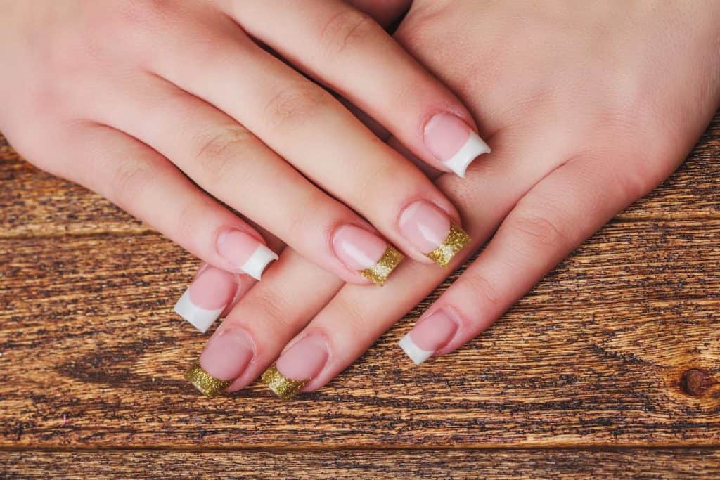 A closeup of a woman's fingernails with a nude nail polish base that has white French tips and gold French tips
