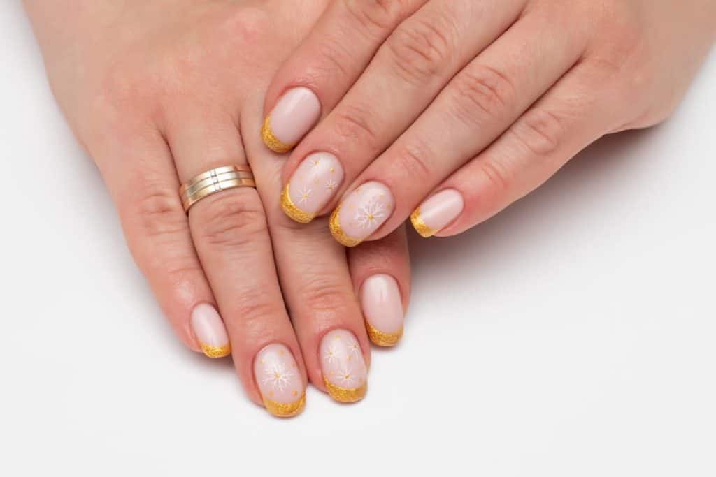 A woman's fingernails with a nude nail polish base that has gold french tips and white snowflakes on select nails