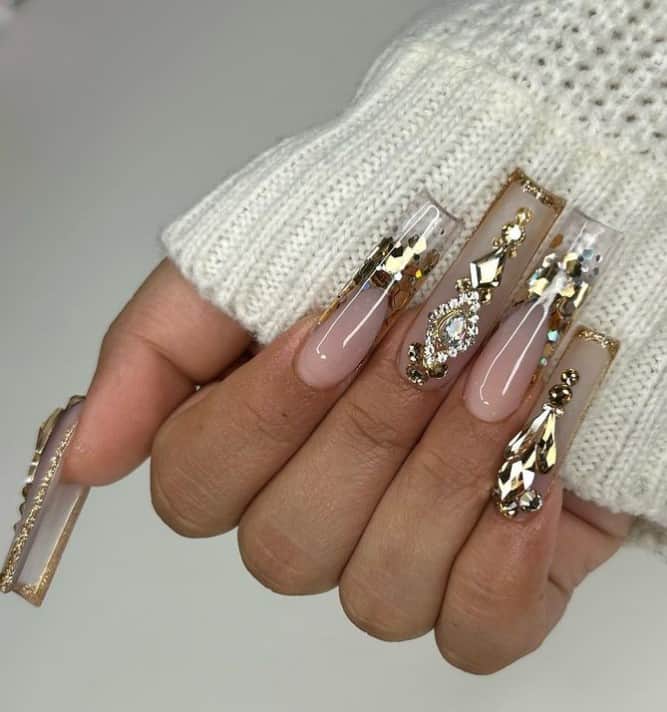 A closeup of a woman's fingernails with a nude to clear nail polish base that has gold tips and clear tips with oversized sequins and large rhinestones