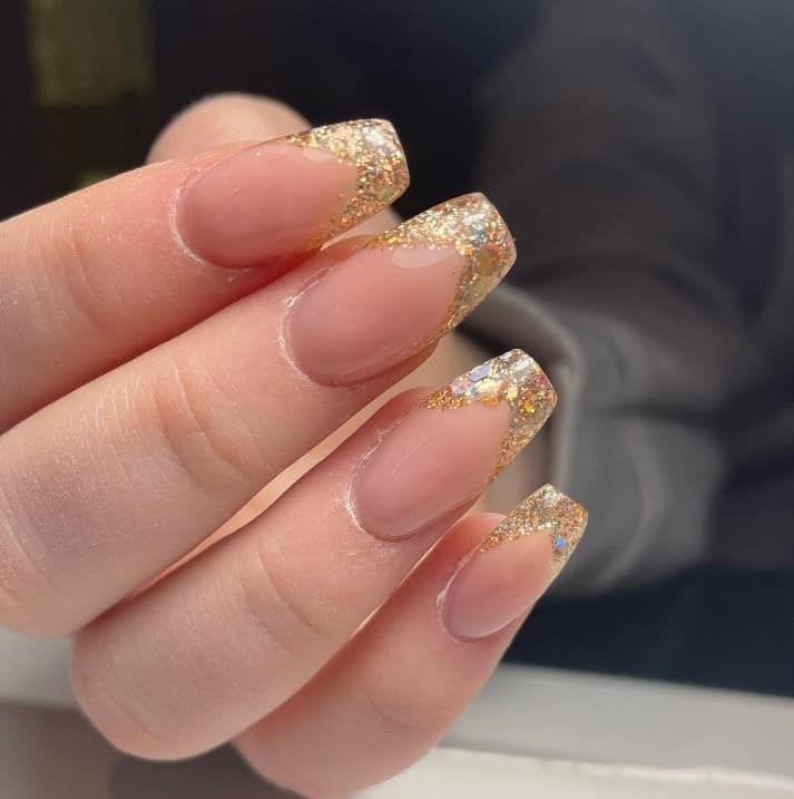 A closeup of a woman's medium-length fingernails with a glossy nude nail polish base that has thick gold glitter French tips and large iridescent glitter flakes