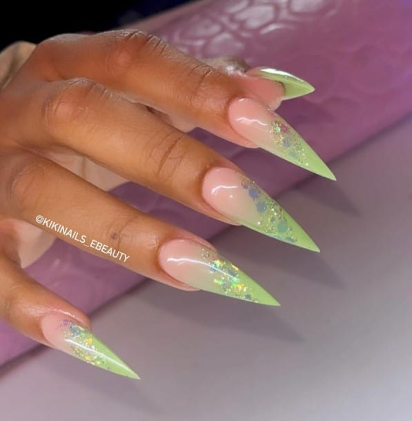 A closeup of a woman's fingernails with a nude and light green ombré nail polish that has small and large glitter flakes in a diagonal line