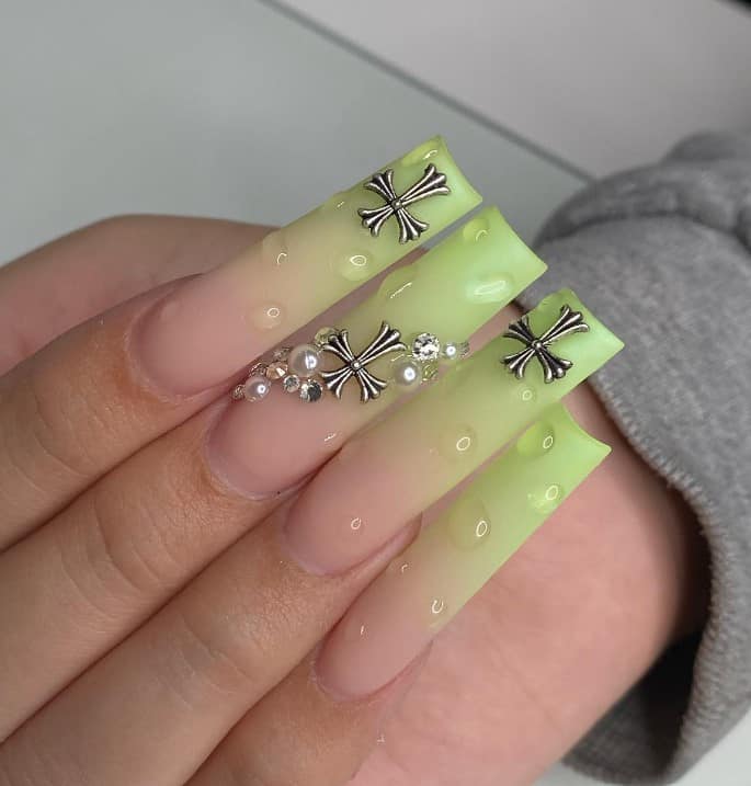 A closeup of a woman's fingernails with a light-sage-green-and-soft-peach ombré nail polish that has translucent droplets, pearls, and silver crosses