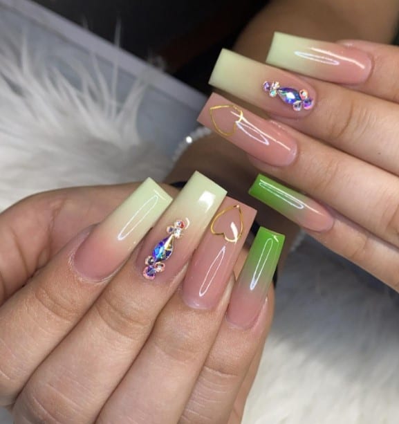 A closeup of a woman's fingernails with earthy green and warm beige nail polish that has white ombré nails, gold hearts and rhinestones on select nails 
