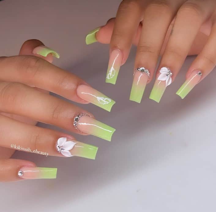 A woman's fingernails with a nude to pastel green gradient effect nail polish that has white petals and silver gems