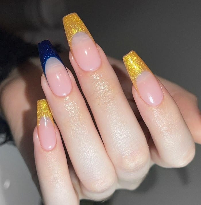 A closeup of a woman's fingernails with a glossy nude nail polish that has shimmery blue tips and shimmery gold tips 