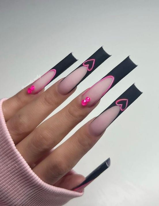 A woman's fingernails with a pale pink nail polish that has black tips, pink heart and pink studs nail designs