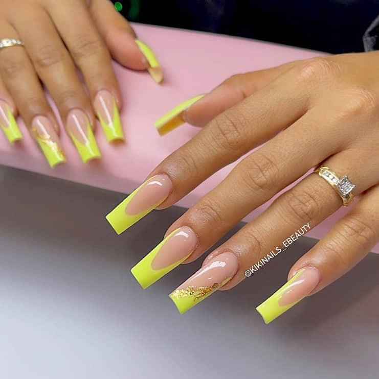 A closeup of a woman's fingernails with a nude nail polish that has bright shade of yellow nail tips and gold foil flakes on select nails