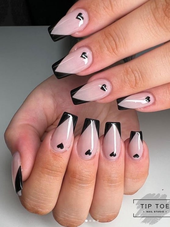 A closeup of a woman's fingernails with a nude nail polish base that has black V-tips and little black hearts