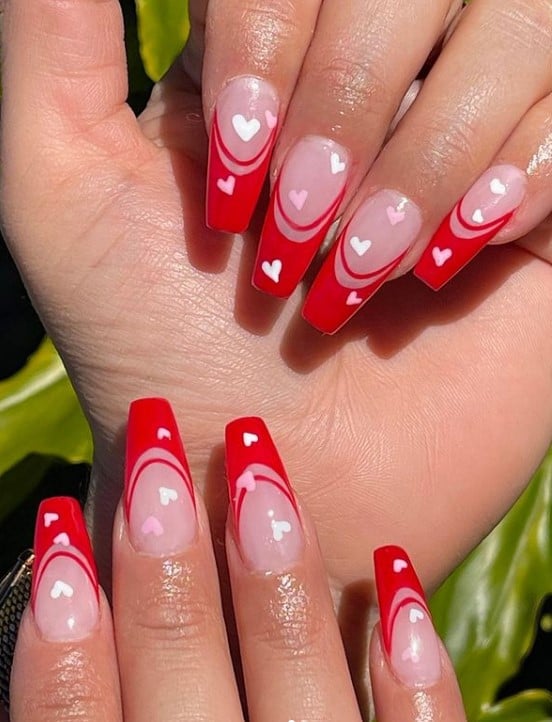A closeup of a woman's fingernails with solid red tips and thin red curve under that has pink and white hearts on top