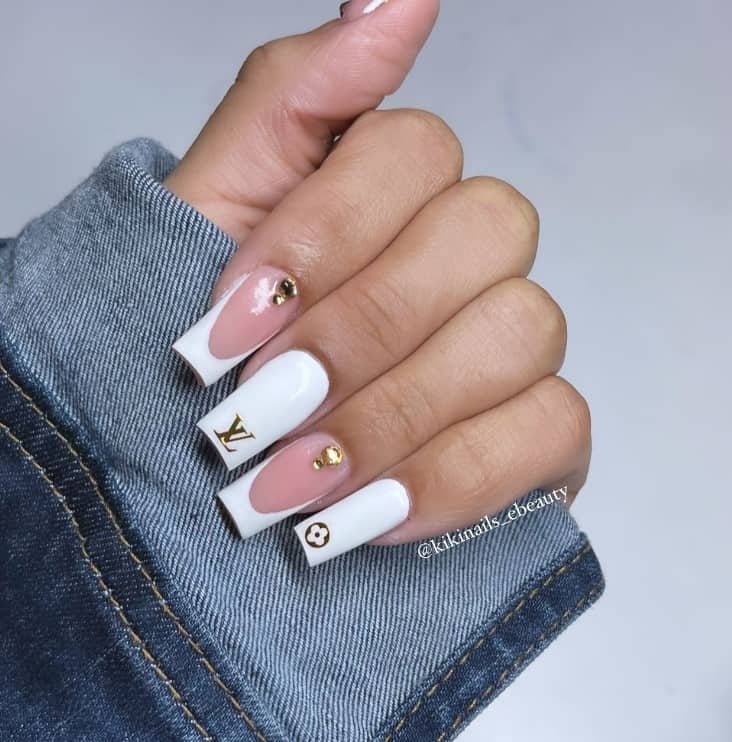 A closeup of a woman's fingernails with a combination of white and nude nail polish that has alternating gold rhinestones and gold LV symbols
