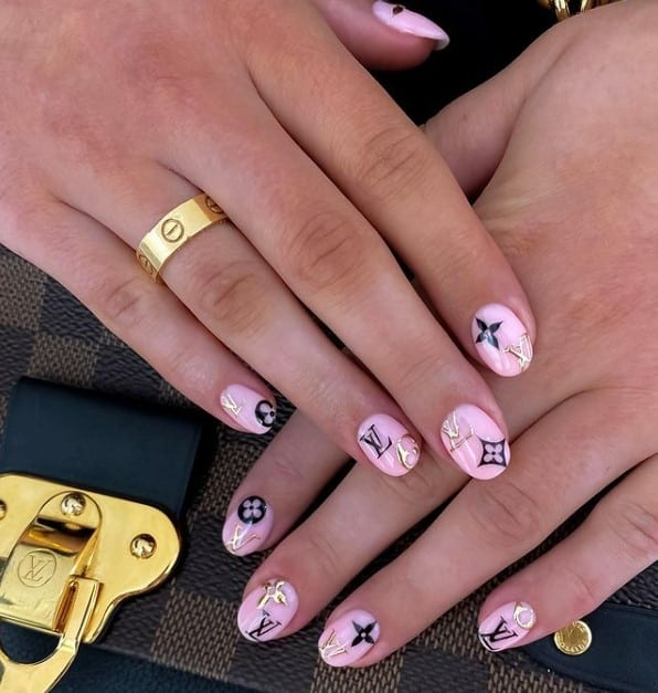 A woman's fingernails with light-pink nail polish base that has black and gold Louis Vuitton nail art