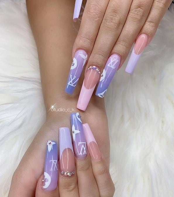 A woman's long coffin fingernails with a purple ombré nail polish that has a sprinkling of LV logo, baby pink and lavender French tips with rhinestones