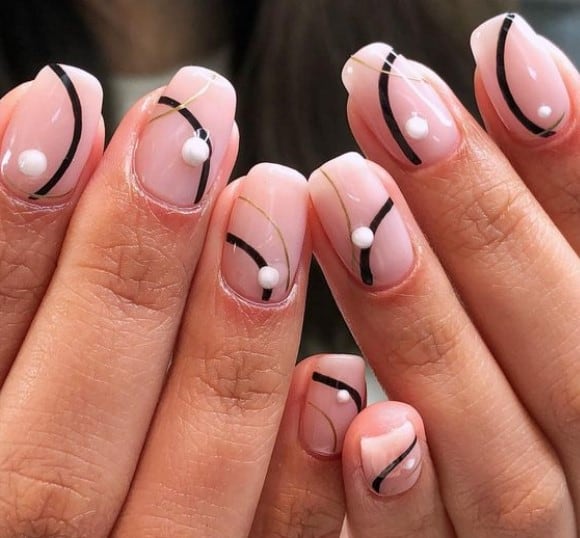 A closeup of a woman's fingernails with sheer pinkish-nude nail polish that has overlapping gold and black curved lines