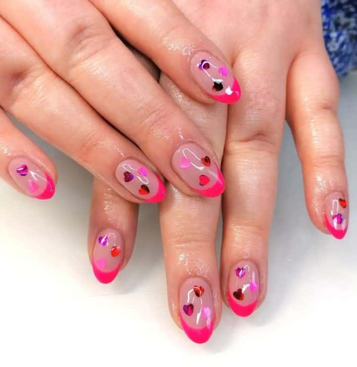 A closeup of a woman's fingernails with a nude nail polish base that has neon pink French tips and multicolored hearts