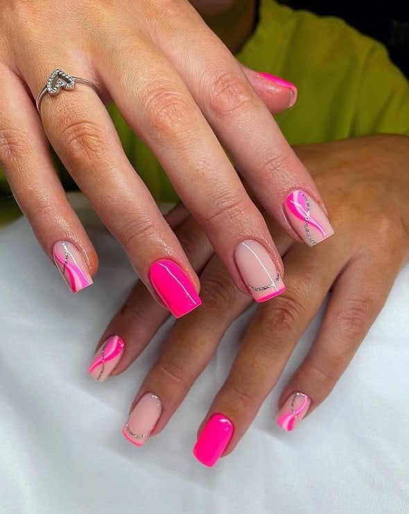 A woman's fingernails with a combination of nude and neon pink nail polish that has three hues of pink with swirls and silver glitter on select nails 