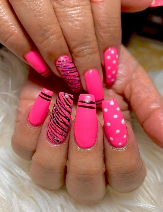 A closeup of a woman's fingernails with a hot pink nail polish that has black glitter tiger stripes and white polka dots