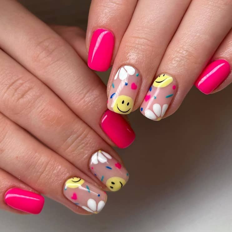 A closeup of a woman's fingernails with a nude and neon pink nail polish base that has bright smileys, flowers, hearts, and sprinkles