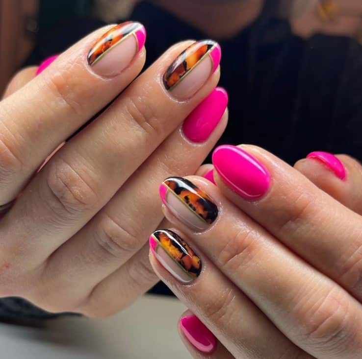 A closeup of a woman's fingernails with a nude and neon pink nail polish that has tortoiseshell design and gold outlines