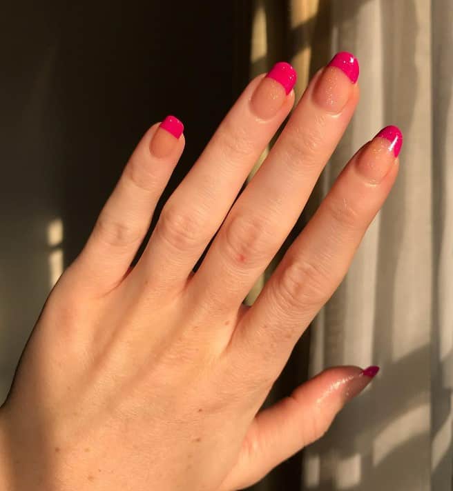 A closeup of a woman's fingernail with French tips bright and obvious that has a nude set of nails with a subtle glitter finish in thick bands of neon pink at the tips