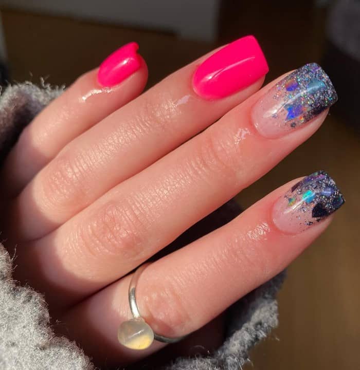 A closeup of a woman's fingernail with neon pink nails that has nails featuring glittery blues, black, silver, and stars