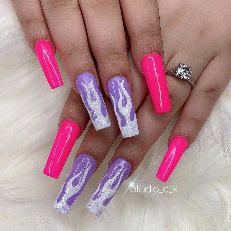 A closeup of a woman's acrylic nail design fingernails with neon pink nails and purple periwinkle accent nails that has white flames and sprinkled with silvery glitter