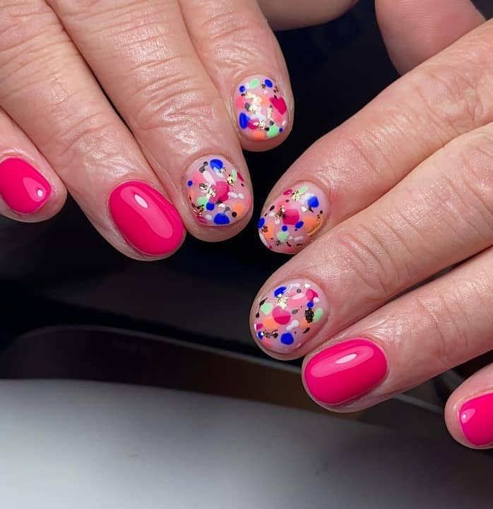 A closeup of a woman's fingernails with short and round pink nails that has paint splatters in multiple colors on nude accent nails