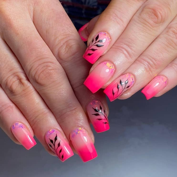 A closeup of a woman's fingernails with glossy neon pink ombre nails that has plant silhouettes and lavender and gold chunky glitter at the cuticles