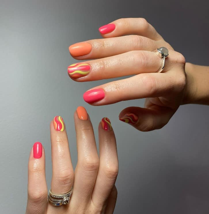 A woman's fingernails with a combination of pink and orange nail polish that has yellow swirls on accent nails 