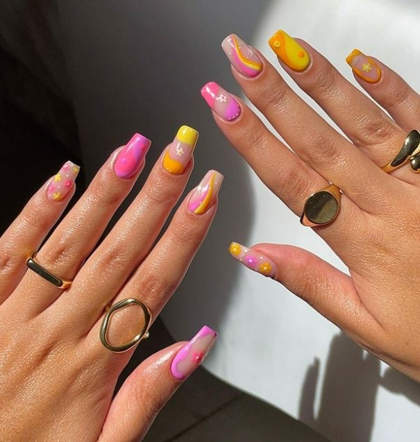A woman's fingernails with a multicolored nail polish that has tiny flowers, yin-yang art, swirls, traditional and inverted French tips, and wavy outlines using bright yellow, pink, and orange nail polish