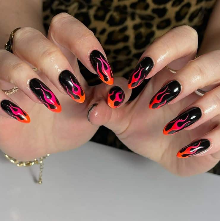 A woman's fingernails with a black nail polish that has pink-and-orange flames on nail tips 