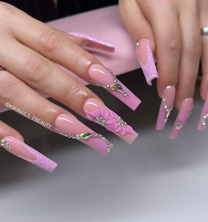 A closeup of a woman's fingernails with a nude pink nail polish base that has 3D flowers, sparkling diamonds and pink glitter at the tip