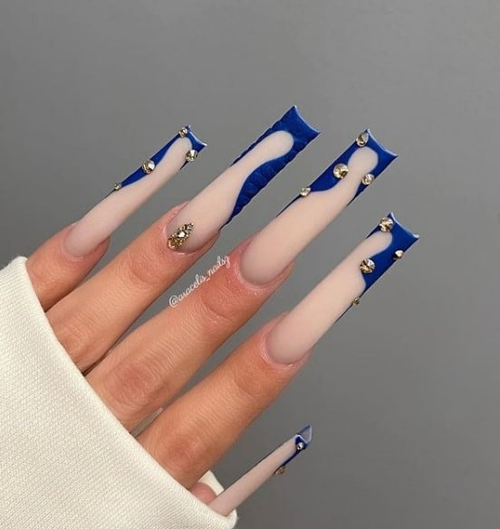 A woman's fingernails with a nude nail polish that has rhinestones, silver rhinestones and wavy royal blue French tips