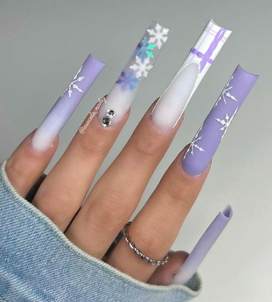 A closeup of a woman's fingernails with a combination of purple and white nail polish that has snowflake nail art, purple plaid French tips, sparkling snowflake stickers, and lovely rhinestones