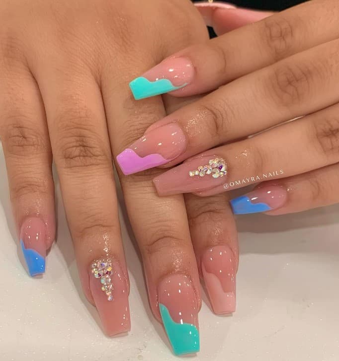 A closeup of a woman's fingernails with a glossy nude nail polish that has multicolored wavy diagonal nail tips and rhinestones on select nails 