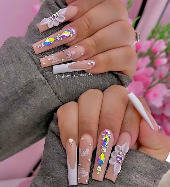 A woman's fingernails with a nude nail polish base that has butterflies, stunning 3D flowers, chic white French tips, and rainbow-colored rhinestones