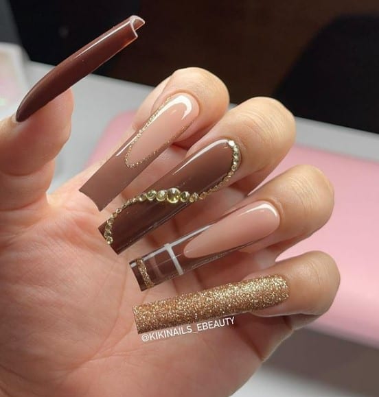 A closeup of a woman's fingernails with a combination of nude, brown and gold glitter nail polish that has sparkling round rhinestones