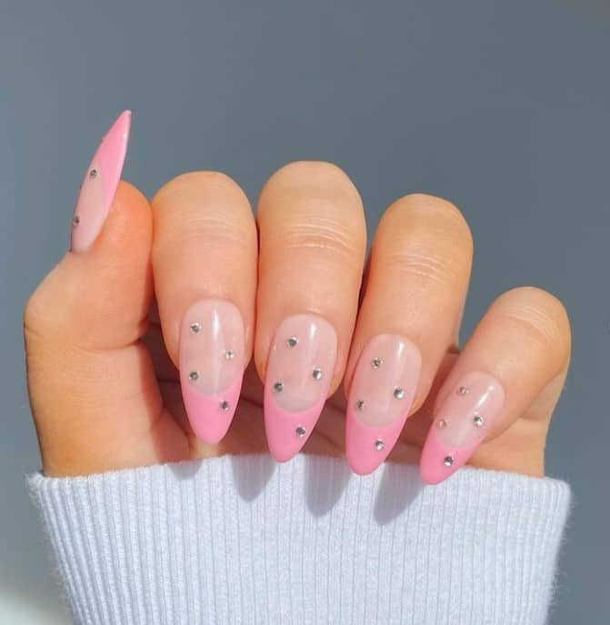 A closeup of a woman's almond-shaped fingernails with a nude nail polish base that has pink French tips and polka-dot rhinestones