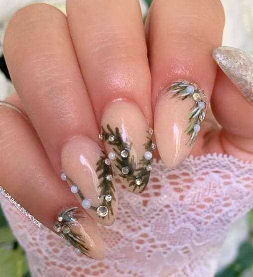 A closeup of a woman's fingernails with a nude nail polish base that has leaf designs and white and clear rhinestones